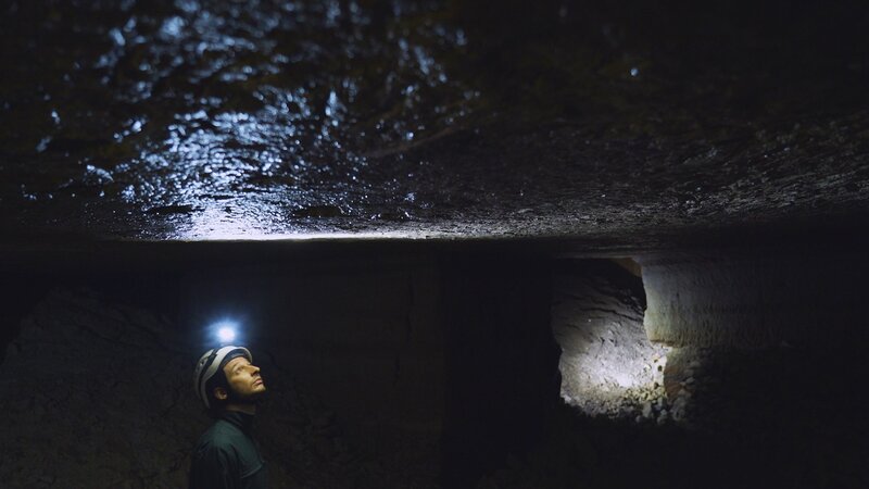 Das Bild zeigt einen unterirdischen Raum mit niedriger Decke und groben Steinwänden, beleuchtet durch eine Lampe, die an einem Helm befestigt ist. An der Decke ist eine feuchte Oberfläche zu sehen, die das Licht reflektiert, was auf das Vorhandensein von Feuchtigkeit oder Wasser hinweist. Wände und Decke wirken wie natürlicher oder von Hand bearbeiteter Stein, was typisch für Katakomben ist. – Bild: PERNEL MEDIA, THE HISTORY CHANNEL