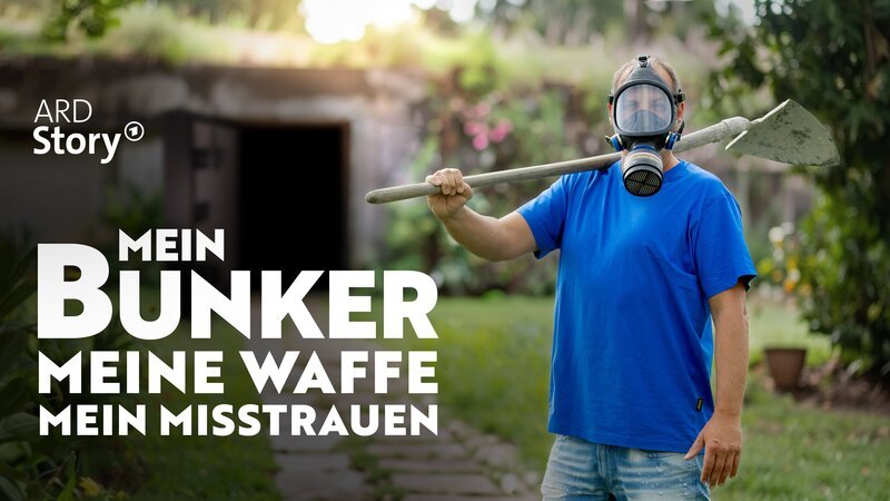 Keyvisual: Christian Klaus mit Gasmaske trägt einen Spaten über der Schulter, er steht in einem Garten, im Hintergrund ist der offene Eingang eines Bunkers zu sehen. – Bild: Katrin Rohde /​ BR /​ NDR /​ MDR /​ Nordend Film Hamburg