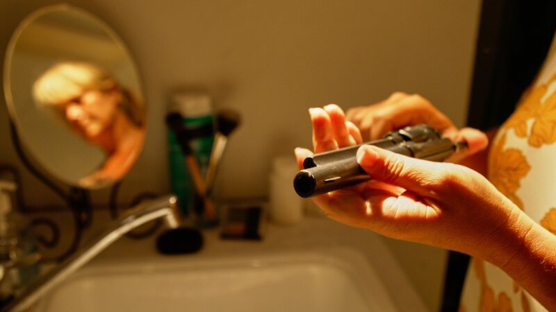 In einem Badezimmer hält eine Person eine Pistole in den Händen, während ihr unscharfes Spiegelbild im Hintergrund erscheint. – Bild: Crime + Investigation