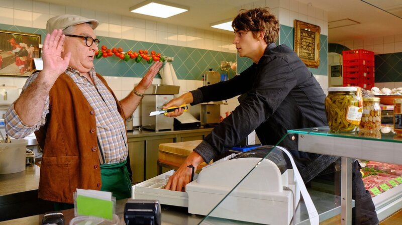 Klaus Scheffler ist in seinem Lebensmittelgeschäft zugange, als ein junger Mann ihn mit einem Teppichmesser bedroht. – Bild: MDR/​Saxonia Media/​Sebastian Kiss