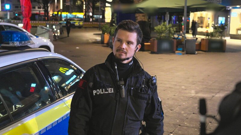 Lukas Weide-Hannemann standing next to a police car looking to the left – Bild: Warner Bros. Discovery, Inc. or its subsidiaries and affiliates.