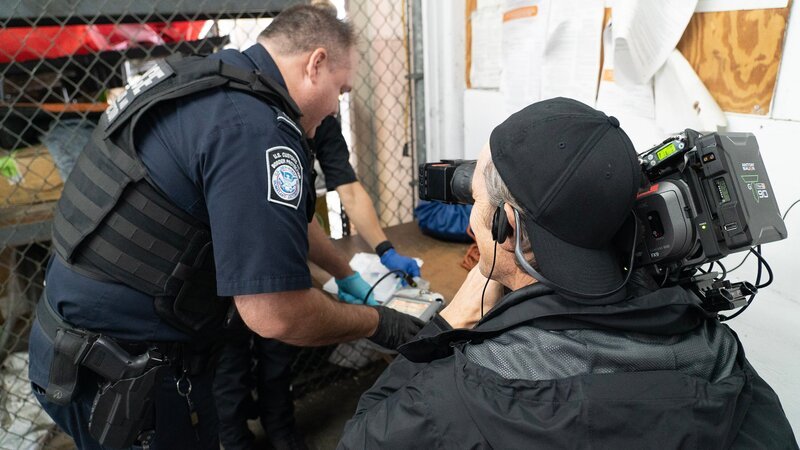 Ein Kameramann filmt den CBP-Beamten Ryan, wie er mit einem Gemini-Gerät den Inhalt einer zerbrochenen Styroporkühlbox untersucht, die auf der Ladefläche eines Lastwagens gefunden wurde, der über den JFK-Flughafen in New York City, N.Y., verschifft wurde (National Geographic for Disney) – Bild: National Geographic for Disney /​ Disney