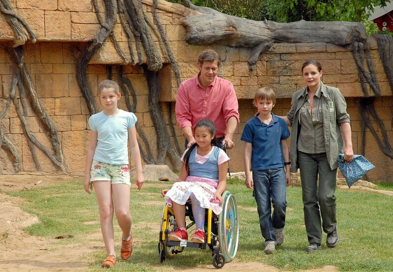 Susanne Mertens (Elisabeth Lanz, rechts) und Christoph Lentz (Sven Martinek, Mitte) mit den Kindern Jonas Mertens (Ludwig Zimmeck, 2. von rechts) und Rebecca Lentz (Elisabeth Böhm, links) und mit Chea San (Linh Pham Dan, im Rollstuhl) auf dem Weg zur Elefantenanlage: Sie wollen das kambodschanische Mädchen dazu überreden, erste Gehversuche mit ihrer Beinprothese zu machen. – Bild: MDR/​Christa Köfer