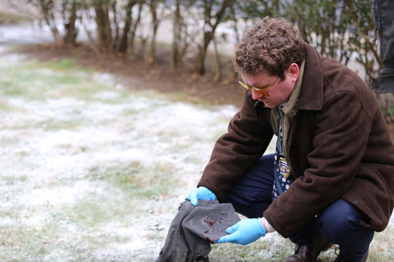 Reconstruction Actor playing Det Schnell (Scott Watson) in brown jacket and yellow glasses crouching to investigate a piece of clothing – Bild: Discovery, Inc.
