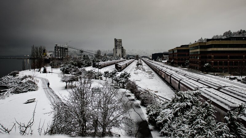Long lines of snow covered train cars wait to be moved following a surprise snow storm. A lone walker follows the trail through Myrtle Edwards Park in Seattle. – Bild: Brian Xavier