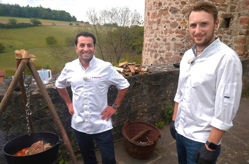 Eine neue Herausforderung für den Münchner Gourmetkoch Ali Güngörmüs. Sein Berufskollege Moritz Zinn (re.) hat ihn nach Homberg/​Efze in sein Restaurant am Fuße der Burg Wallenstein eingeladen. Der junge Outdoor-Küchen-Fan will Ali mit einem uralten Familienrezept überzeugen: Omas Lammtopf – gekocht über dem offenen Feuer. – Bild: HR/​Sabine Guth