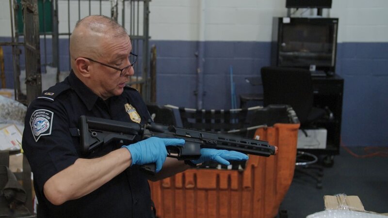 CBP-Beamter Cisneros hält eine Waffe in der Hand, die in einem verschickten Paket in der Poststelle des JFK International Airport gefunden wurde, während er erklärt, warum eine orangefarbene Spitze an gefälschten Feuerwaffen angebracht sein muss. – Bild: Courtesy of Disney+