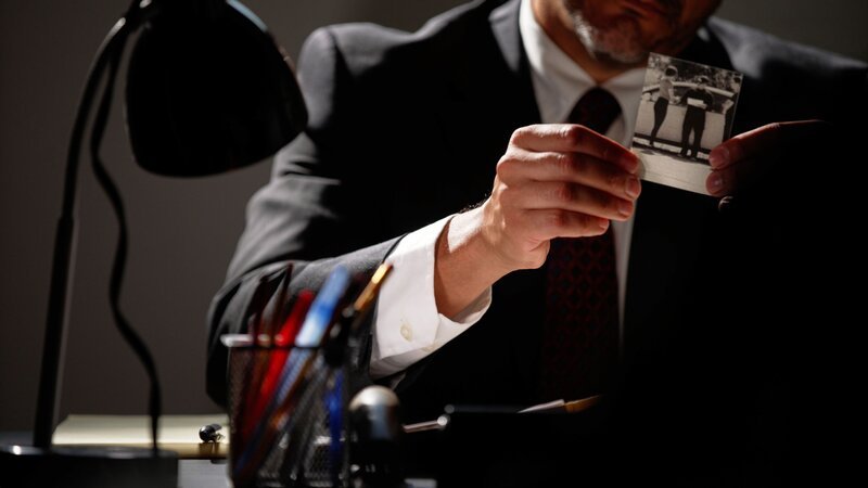 Eine Person in formeller Kleidung sitzt an einem Schreibtisch und hält eine Schwarz-Weiß-Fotografie. Auf dem Schreibtisch befinden sich Büroartikel wie Stifte und eine Lampe, was auf eine professionelle oder ermittelnde Umgebung hinweist. – Bild: Crime + Investigation