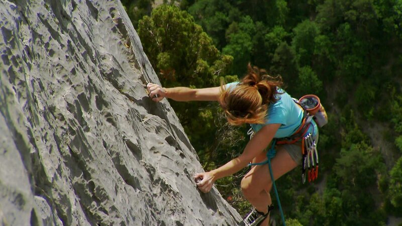 Ein Kletterer erklimmt eine steile Felswand, gesichert mit Seilen und Kletterausrüstung. Die raue Steinoberfläche steht im Kontrast zum dichten grünen Wald darunter und erzeugt eine eindrucksvolle Outdoor-Szenerie. – Bild: NDR