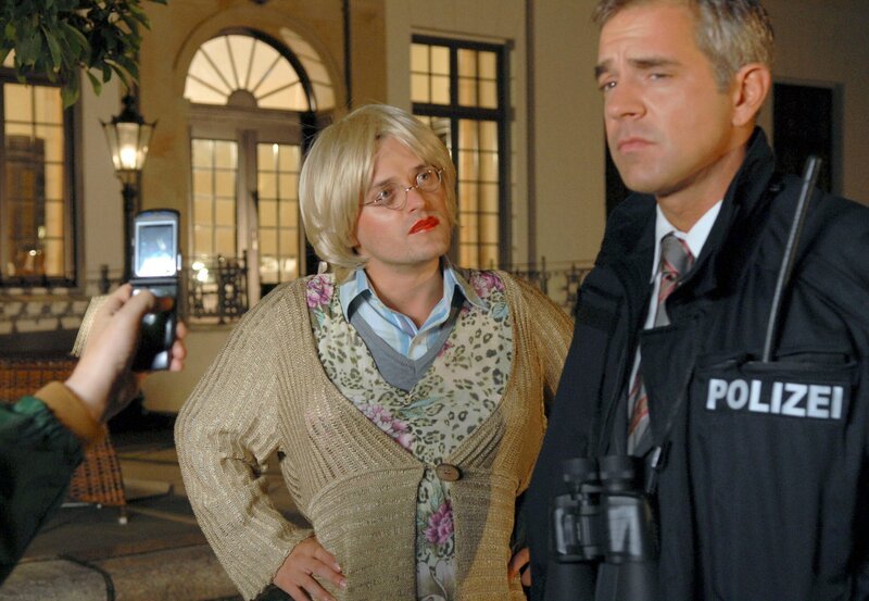 MDR Fernsehen ELVIS UND DER KOMMISSAR I. STAFFEL FOLGE 6, „Heiße Ware“, am Donnerstag (24.06.10) um 08:40 Uhr. Dieser Großeinsatz ist mehr als ein Foto wert: Assistent Pröttel (Stefan Haschke, M.) fungiert dabei als Double für Rosa Behringer, trägt Rouge und üppige Frauenkleider – und wird per Handykamera geknipst. Sein Chef Seibold (Florian Fitz, r.) sieht auch mit Polizeijacke nicht professioneller aus. Kommissar Behringer ermittelt derweil an einem ganz anderen Ort. – Bild: MDR/​Marion von der Mehden