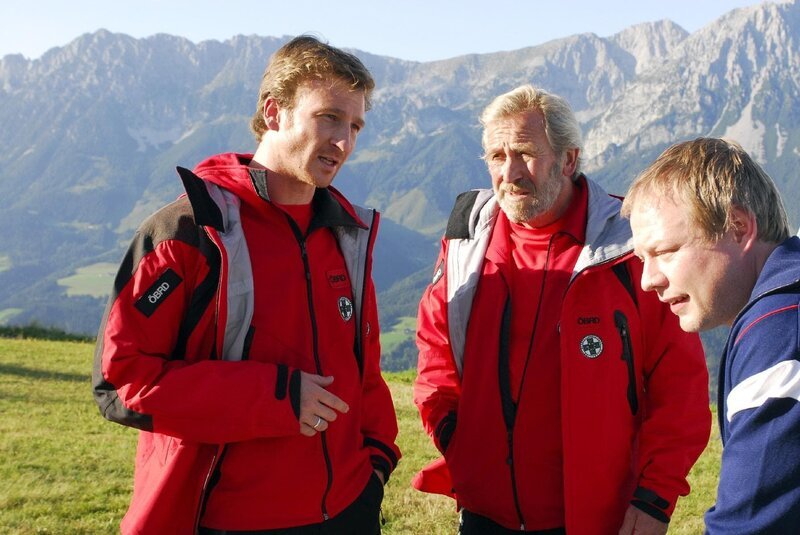 Hans (Heiko Ruprecht, li.) und Franz (Hermann Giefer, mi.), werden vom Fallschirmverleiher (Wowo Habdank, re.) darüber informiert, dass zwei seiner Kunden von ihrem Flug nicht zurück sind. Als Hans erkennt, dass es sich dabei um Martin und Andrea handeln muss, ahnt er nicht, dass die beiden bereits um ihr Überleben kämpfen. – Bild: ORF/​ZDF/​Hans Seidenabel