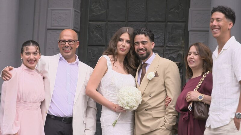 The Bride Kathrin and the groom Yassir are standing between Yassir’s parents and siblings and smiling to the camera. Kathrin is holding a white flower bouquet. – Bild: Warner Bros. Discovery, Inc. or its subsidiaries and affiliates.