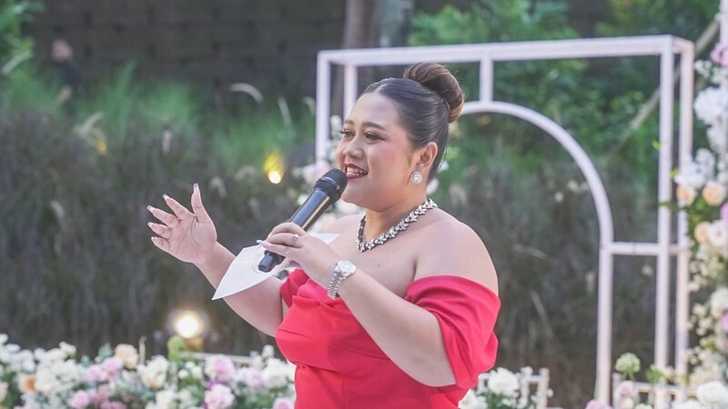 Wedding Planner Julie is holding a microphone in her hand as well as presentation cards. She is wearing a read dress and her hair is styled to a formal bun. – Bild: Warner Bros. Discovery, Inc. or its subsidiaries and affiliates. All rights reserved.