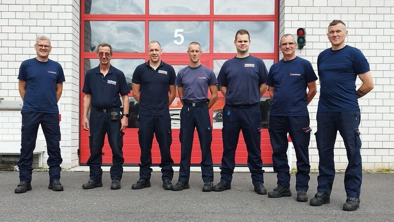 Seven firefighters in front of the vehicle depot in Fürstenwalde/​Spree. – Bild: Warner Bros. Discovery, Inc. or its subsidiaries and affiliates