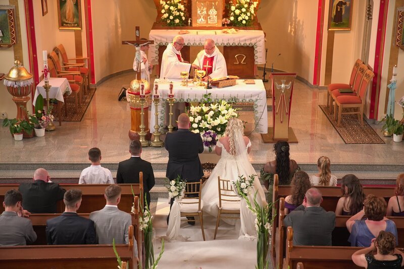 Photograph from the church. The priests are standing behind the altar, the bridal couple is in front of them. The wedding guest are kneeing on the church benches. – Bild: Production Company /​ Warner Bros. Discovery, Inc.