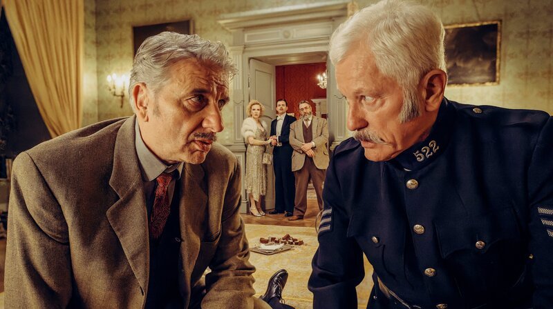 Chief Inspector Francis Lightmyer (Udo Wachtveitl, links vorne) und Constable Ivor Partridge (Miroslav Nemec, rechts vorne) untersuchen Arthur im Kaminzimmer. Lady Bantam (Sunnyi Melles), Charles (Ferdinand Hofer) und Dr. Mallard (Alexander Hörbe, rechts hinten) beobachten sie. – Bild: BR/​Bavaria Fiction GmbH/​Hendrik Heiden