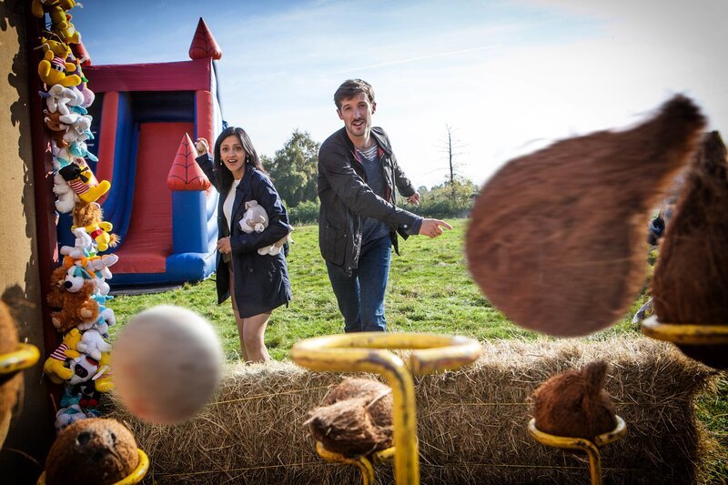 Charlie Nelson (Gwilym Lee, r.) und die Gerichtsmedizinerin Kam Karimore (Manjinder Virk, l.) vergnügen sich auf einem Jahrmarkt beim Werfen nach Kokosnüssen. – Bild: ZDF/​Mark Bourdillon 07831605033