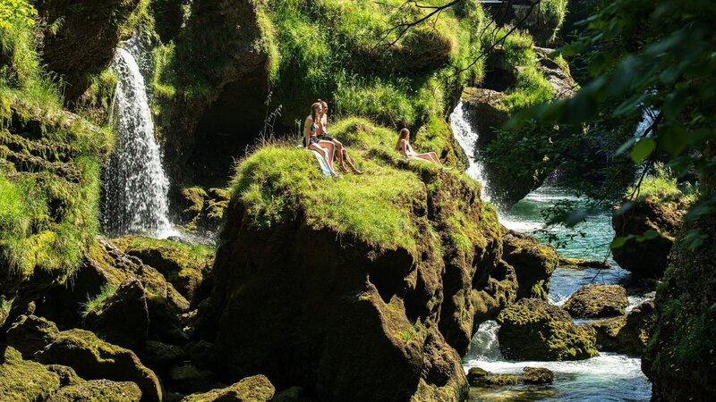 Menschen sitzen auf einem sonnenbeschienenen, grasbewachsenen Felsen neben kleinen Wasserfällen über einem klaren Fluss. – Bild: @Red Bull Media House/​ServusTV