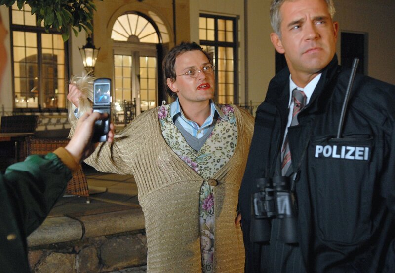 Dieser Großeinsatz ist mehr als ein Foto wert: Assistent Pröttel (Stefan Haschke, M.) fungiert dabei als Double für Rosa Behringer, trägt Rouge und üppige Frauenkleider – und wird per Handykamera geknipst, als er sich die Perücke vom Kopf reißt. Sein Chef Seibold (Florian Fitz, r.) sieht auch mit Polizeijacke nicht professioneller aus. Kommissar Behringer ermittelt derweil an einem ganz anderen Ort. – Bild: NDR/​ARD/​Marion von der Mehden
