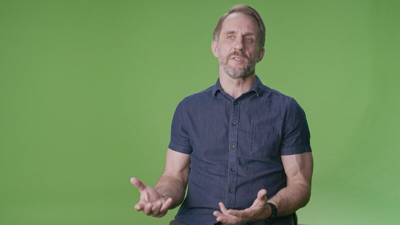 Eine Person sitzt vor einem einfarbigen grünen Hintergrund und trägt ein kurzärmeliges, dunkelblaues Hemd mit Knöpfen. Die Hände sind geöffnet und gestikulieren, was auf einen erklärenden oder Gesprächsmoment in einem Studio hinweist. – Bild: A+E Global Media /​ THE HISTORY CHANNEL