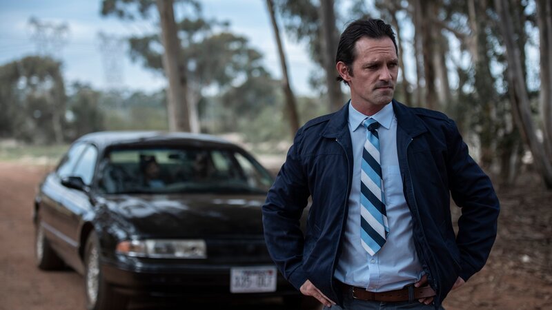 Drama: A detective stands near his car in a wooded area, looking pensive. – Bild: Anika Molnár /​ Discovery Communications, LLC