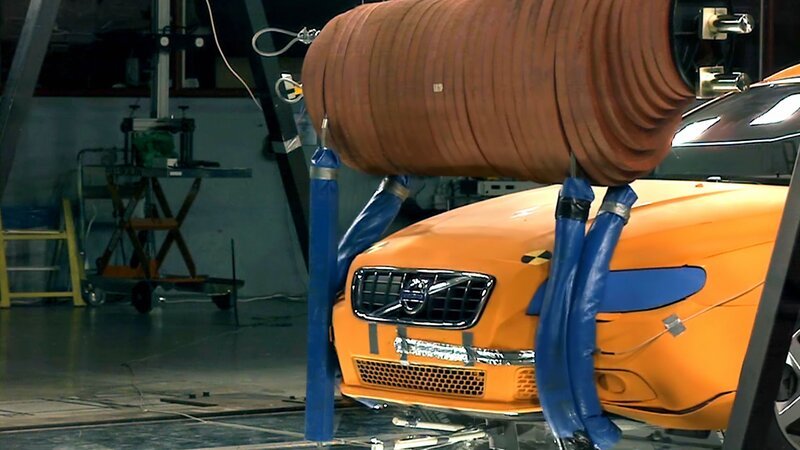 Ein leuchtend orangefarbenes Volvo-Fahrzeug befindet sich in einer Crashtest-Anlage, wobei ein großer zylindrischer Aufprallkörper über der Motorhaube hängt. Das Auto ist auf einer Prüfplattform fixiert, und blaue Schutzpolster umgeben die Haltearme des Aufprallkörpers. Die Szene zeigt industrielle Geräte und Kabel, was auf eine kontrollierte Umgebung für Fahrzeugsicherheitsprüfungen hinweist. – Bild: ZDF /​ VOLVO /​ THE HISTORY CHANNEL