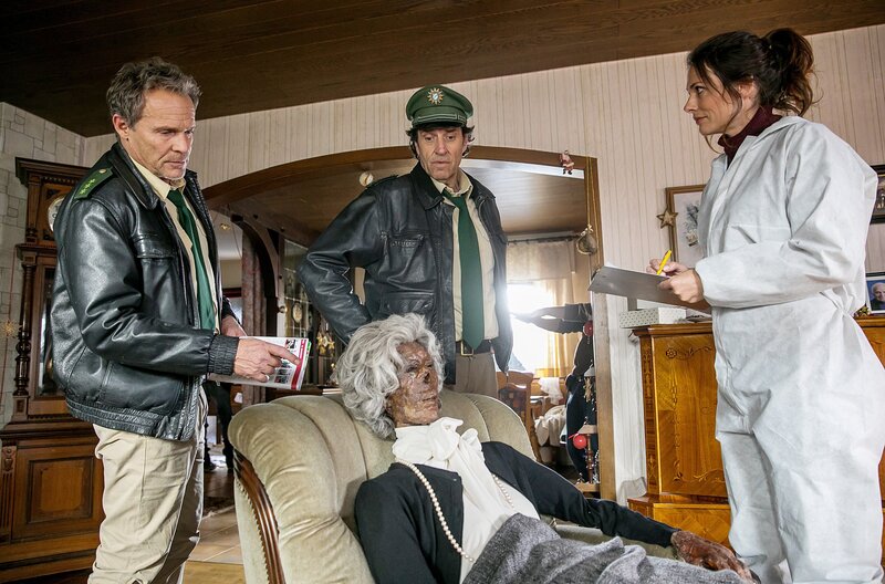 Hubert (Christian Tramitz) und Staller (Helmfried von Lüttichau) finden die Seniorin mumifiziert in einem Sessel sitzend; die Pathologin Dr. Fuchs (Susu Padotzke) geht zunächst von einem natürlichen Tod aus. – Bild: ARD/​TMG/​Chris Hirschhaeuser