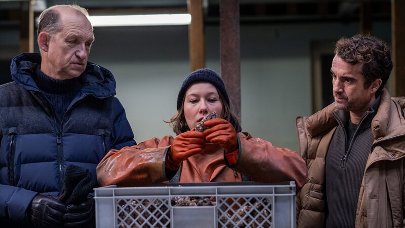 Tomke Bundis (Josefine Israel, M.) bei der Arbeit. Die Austernbänke bei denen Wolf Voss gefunden wurde gehören ihr, doch mit seinem Tod will sie nichts zu tun gehabt haben. Immer wieder hat sich der Gastronom an ihren Austern bedient, doch dafür bringt sie doch niemandem um! Die Kommissare Carl Sievers (Peter Heinrich Brix, l.) und Hinnerk Feldmann (Oliver Wnuk, r.) sind nicht überzeugt. – Bild: ZDF und Manju Sawhney