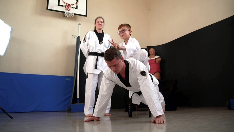 Bei diesem Prank wird Taekwondo-Trainer Nils (M.) an seine Grenzen gebracht. Los geht es mit Liegestützen, erschwert durch den 9-jährigen Taylan (r.), der fröhlich auf seinem Rücken sitzt. Trainerin Pia (l.) ist gespannt. – Bild: ZDF/​Contentladen