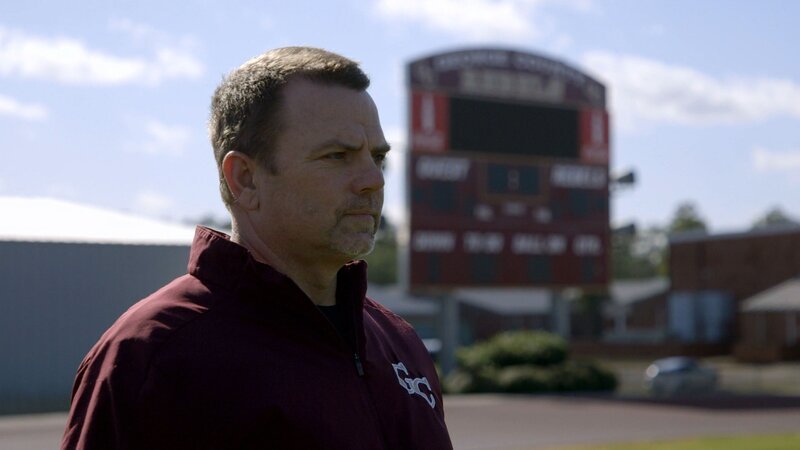 Eine Person in einer dunklen Jacke mit einem Logo und Buchstaben auf der linken Brust steht im Freien; im Hintergrund eine große Anzeigetafel auf einem Sportplatz mit Text und Zahlen sowie Gebäuden, die an Schul- oder Sportanlagen erinnern. Klarer Himmel mit einigen Wolken. – Bild: Crime + Investigation /​ A+E Networks