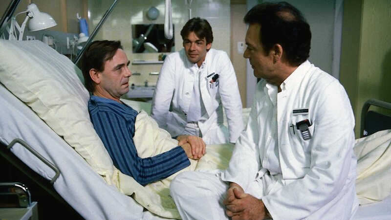 Dr. Rens (Holger Petzold, l.) ist plötzlich erkrankt. Er sieht einer Operation entgegen. Auch ein Arzt kann Angst vor einer Blinddarmoperation haben. Professor Brinkmann (Klausjürgen Wussow, r.) und sein Sohn Udo (Sascha Hehn, M.) versuchen ihm gut zuzusprechen. – Bild: Thomas Waldhelm