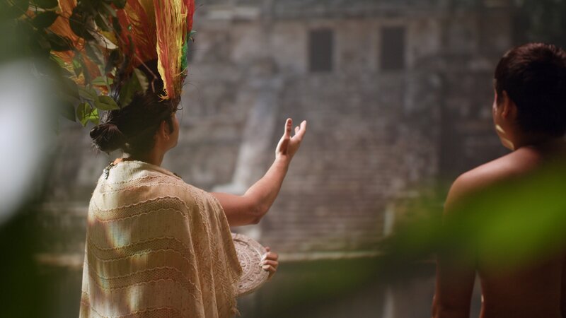 An Ancient Maya timekeeper/​ priest in ceremonial clothing stands at the edge of the sacred cenote holding a stone calendar. – Bild: Go Button Media