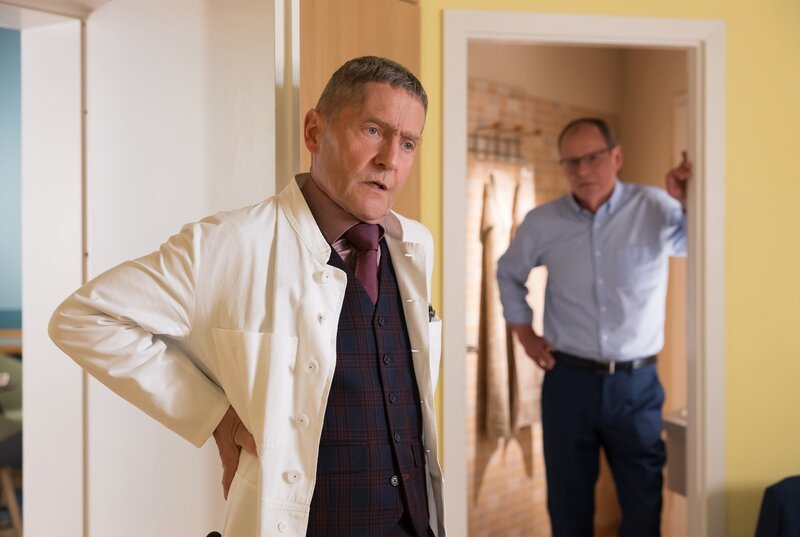 Dr. Harald Reimers (Olaf Burmeister, re.) ist der psychologische Gutachter, der für Dr. Kaminskis (Udo Schenk, li.) MPU zuständig ist. Er bietet dem Arzt einen Deal an. Wenn Kaminski ihn operiert, statt konservativ zu behandeln und er seinen schmerzhaften Nierenstein schneller loswird, steht er gewissermaßen in „Kaminskis Schuld“. Nun ist Kaminski in der Zwickmühle: Mit seinem Hexenschuss, den er vor Reimers verheimlicht, ist er absolut nicht in der Lage eine OP durchzuführen. – Bild: MDR/​Saxonia Media/​Rudolf Wernicke