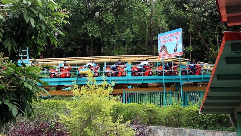 Die Aufnahme zeigt eine moderne Achterbahn in einem Freizeitpark, die durch eine grüne, bewaldete Umgebung verläuft. Die Schienen sind in leuchtendem Blau und Gelb gehalten, und die Wagen sind in einer Motorrad-ähnlichen Form gestaltet, was dem Fahrgeschäft ein dynamisches und innovatives Aussehen verleiht. Mehrere Personen sitzen in den Wagen, während die Bahn in einer geraden Passage fährt. Im Vordergrund sind grüne Pflanzen und Sträucher zu sehen, die die natürliche Atmosphäre des Parks unterstreichen. Ein Schild mit chinesischen Schriftzeichen befindet sich neben der Strecke, was auf den Standort in einem asiatischen Freizeitpark hinweist. – Bild: ZDF /​ CUPCAKES & COASTERS VLOG /​ THE HISTORY CHANNEL