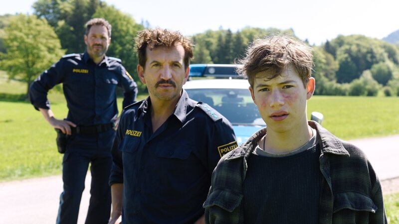 Quentin König (Thilo Fridolin Matthes, r.) berichtet den Polizisten Loisl (Sebastian Gerold, M.) und Charlie (Thomas Birnstiel, l.) aufgeregt von dem Jagdunfall seines Vaters. – Bild: Barbara Bauriedl /​ ZDF