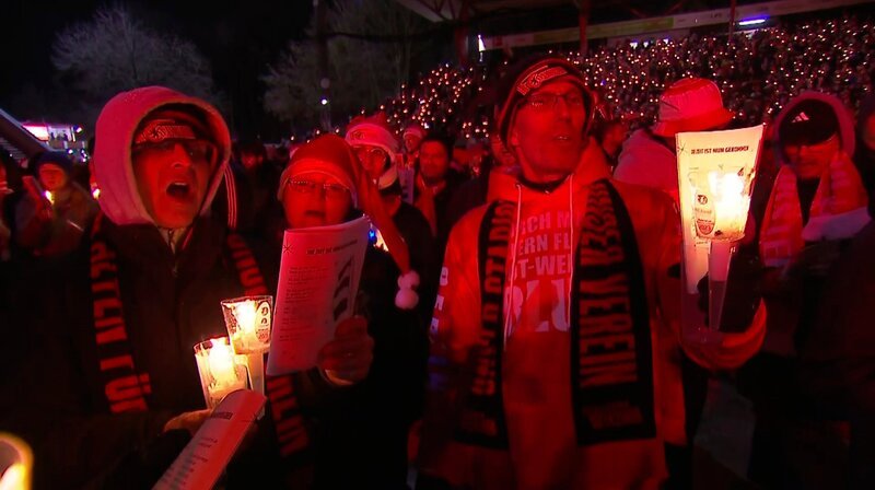 Es war im Jahr 2003, als sich Mitglieder des Fanclubs Alt-Unioner heimlich im Stadion trafen, um nach einer enttäuschenden Hinrunde zum Trost gemeinsam Advents- und Weihnachtslieder zu singen. Mittlerweile kommen jedes Jahr rund 28.500 Fans zusammen und bilden den wohl berühmtesten Weihnachtschor Deutschlands. Wir blicken hinter die Kulissen und stellen die Macher des Weihnachtssingens vor. – Singender Einsatz von echten Union-Fans. – Bild: rbb