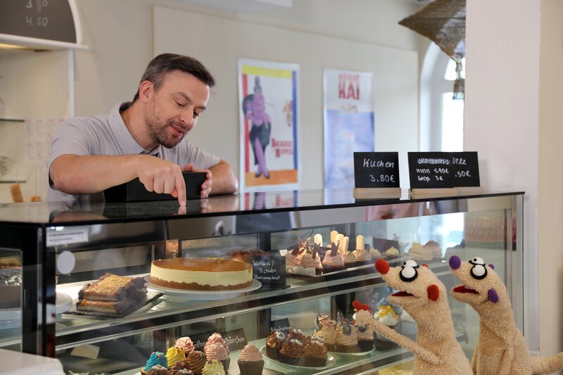Jan und Henry suchen bei einem Konditor (Engin Ben Akkaya) nach Sonnencreme. – Bild: NDR/​bigSmile