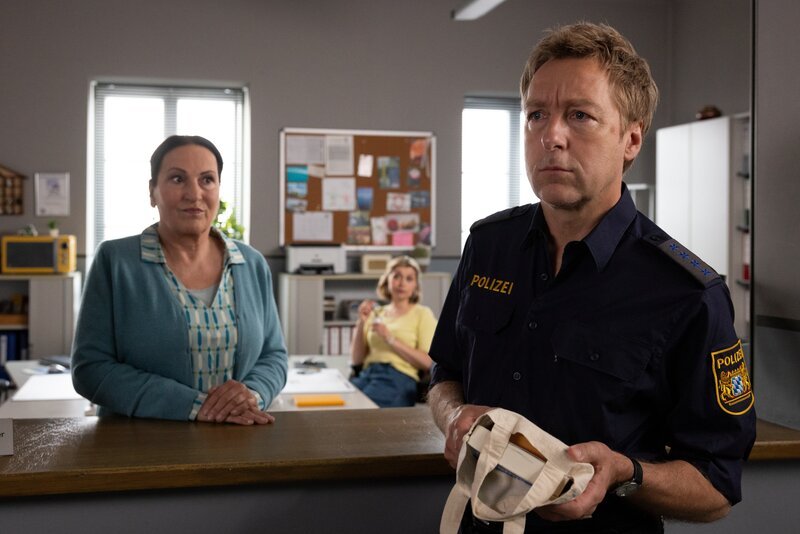 Frau Lange (Sarah Thonig, M.) beobachtet gespannt, wie Frau Grasegger (Ursula Maria Burkhart, l.) versucht, Polizeihauptmeister Mohr (Max Müller, r.) beizubringen, dass die Bücher noch nicht signiert sind. – Bild: ZDF und Linda Gschwentner