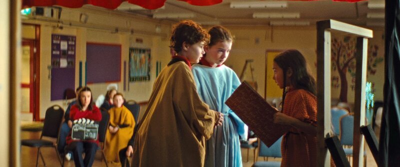 Holly (Portia McLaughlin, rechts), die im Krippenspiel die Rolle der Wirtin ergattert hat, bietet unter dem strengen Blick ihrer Lehrerin (Clare Monnelly, links im Hintergrund) Maria (Maribel Harrison, Mitte) und Josef (James McGowan, links) während der Probe ein Zimmer an. – Bild: KiKA/​Reel Issues Films