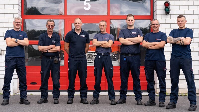Seven firefighters in front of the fire station in Fürstenwalde/​Spree. – Bild: Warner Bros. Discovery, Inc. or its subsidiaries and affiliates. All rights reserved.