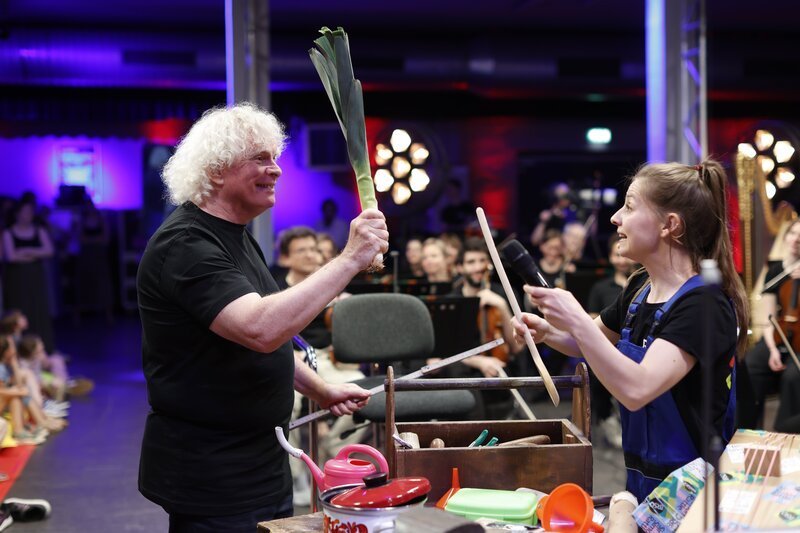Familienkonzerte mit Checkerin Marina und dem BRSO unter der Leitung von Sir Simon Rattle in der TonHalle. Im Bild: Checkerin Marina und Sir Simon probieren verschiedene Objekte als Taktstöcke aus. – Bild: BR/​Astrid Ackermann