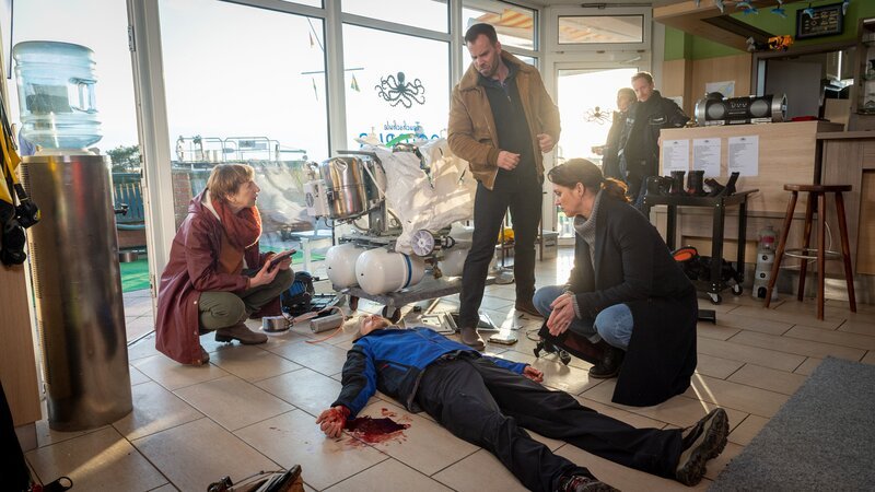 Der Mitinhaber der Wismarer Tauchschule Octopus, Falk Leonhardt (Marcus Kuhn, 2.v.l.), wird erstochen in seinen Geschäftsräumen aufgefunden. Rechtsmedizinerin Dr. Helene Sturbeck (Katharina Blaschke, l.) ist bereits am Tatort und untersucht die Leiche. Die Kommissare Pöhlmann (Dominic Boeer, M.) und Joost (Nike Fuhrmann, r.) sammeln erste Informationen über den Tathergang. Könnte das demolierte Gerät im Schaufenster eine Rolle gespielt haben? Was ist das überhaupt? – Bild: ZDF /​ Meyerbroeker