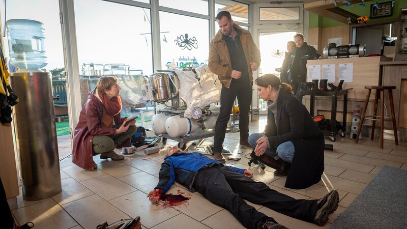 Der Mitinhaber der Wismarer Tauchschule Octopus, Falk Leonhardt (Marcus Kuhn, 2.v.l.), wird erstochen in seinen Geschäftsräumen aufgefunden. Rechtsmedizinerin Dr. Helene Sturbeck (Katharina Blaschke, l.) ist bereits am Tatort und untersucht die Leiche. Die Kommissare Pöhlmann (Dominic Boeer, M.) und Joost (Nike Fuhrmann, r.) sammeln erste Informationen über den Tathergang. Könnte das demolierte Gerät im Schaufenster eine Rolle gespielt haben? Was ist das überhaupt? – Bild: ZDF /​ Meyerbroeker