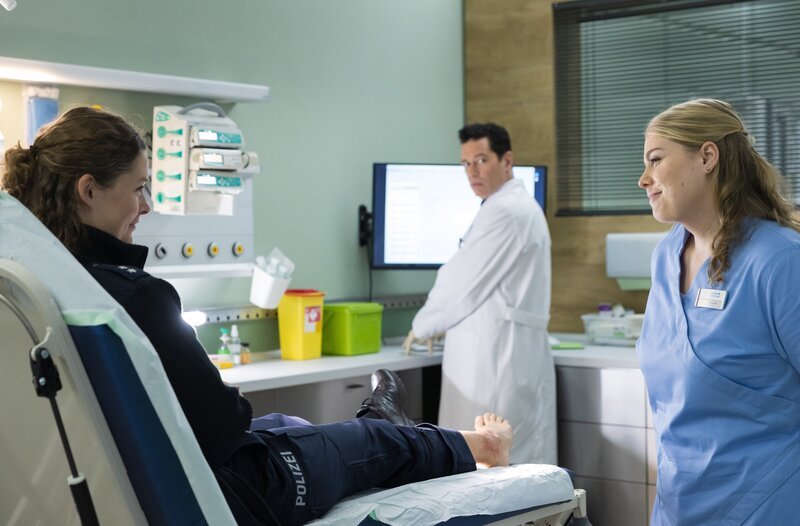 Nele Garbow (Laura Louisa Garde, li.) freut sich, trotz ihres wahrscheinlich gebrochenen Sprunggelenks, dass sie Miriam Schneider (Christina Petersen, re.) so schnell wiedersieht. Dr. Philipp Brentano (Thomas Koch, mi.) ist verwundert, ob der offenen Flirterei der Polizistin. – Bild: Rudolf Wernicke /​ MDR