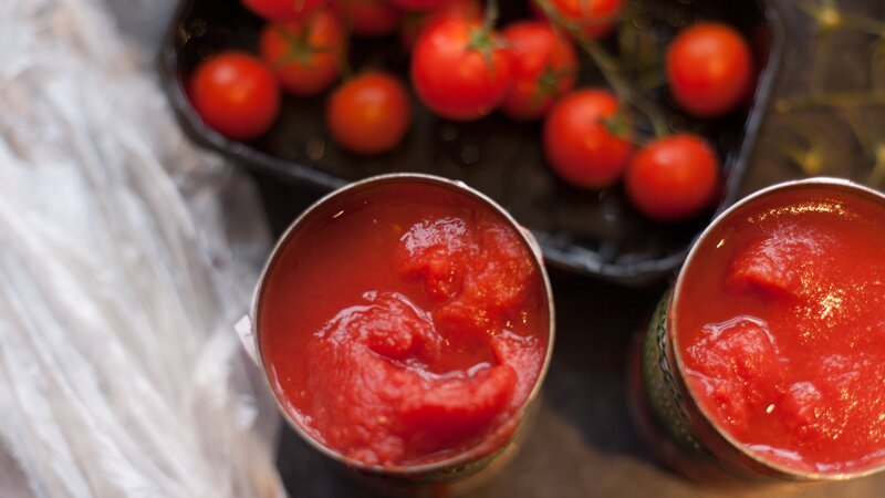 Fresh cherry tomatoes (on the vine) and two open cans of tomatoes on a kitchen counter – Bild: Paula Banks /​ Getty Images