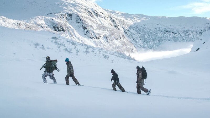 Eine Gruppe von Abenteurern wandert durch eine weite, schneebedeckte Berglandschaft. Umgeben von hohen Gipfeln und tiefen Tälern bewegen sie sich in einer Reihe über das unberührte weiße Gelände und tragen Ausrüstung für ihre Tour in der Wildnis. – Bild: Waidwerk