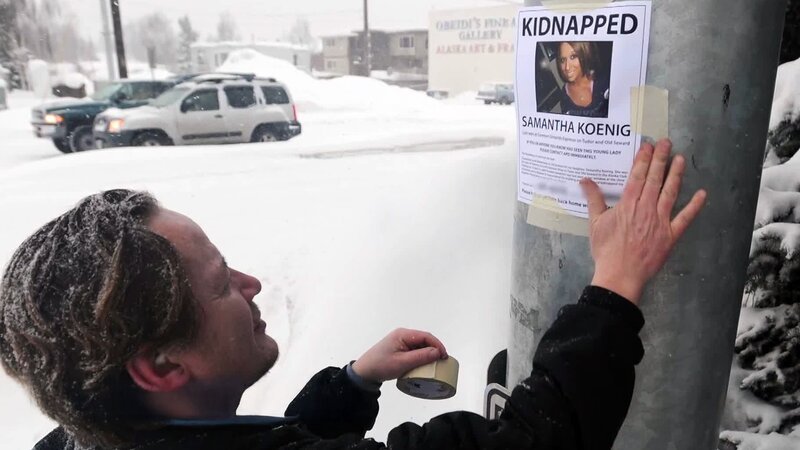 Das Bild zeigt eine Person, die ein Plakat mit Klebeband an einen Metallpfosten befestigt. Auf dem Plakat steht „KIDNAPPED“ und der Name „Samantha Koenig“, zusammen mit einem Foto und Text mit Details zum Verschwinden. Im Hintergrund ist eine verschneite Straße mit geparkten Fahrzeugen und Gebäuden zu sehen. – Bild: A+E Global Media /​ Crime + Investigation