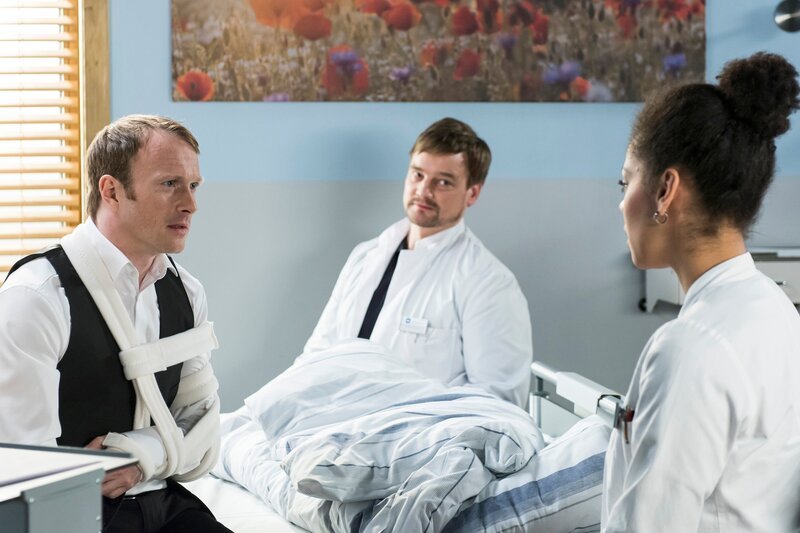 Vivi (Jane Chirwa, r.) muss Bernd Müller (Merten Schroedte, l.) im Beisein von Dr. Lindner (Christian Beermann, M.) mitteilen, dass er einen Tumor in der Schulter hat. – Bild: ARD/​Jens Ulrich Koch
