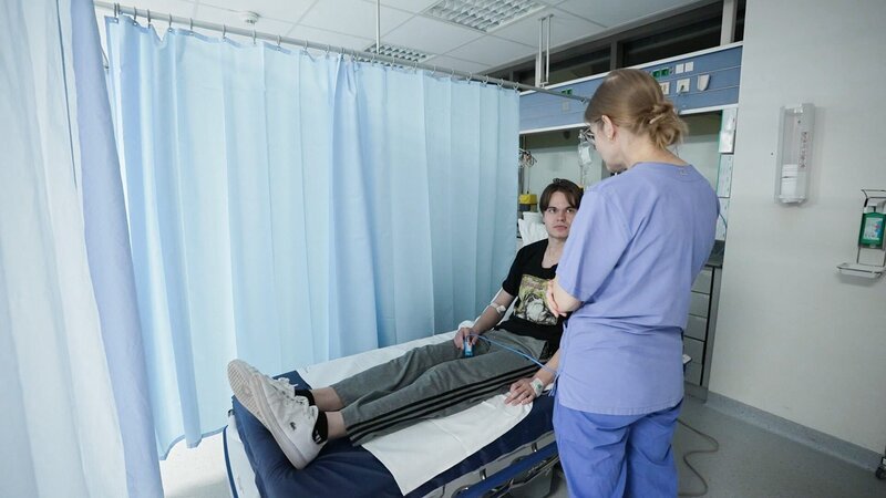Ep5 – A female phyisician wearing blue scrubs and an low bun stands close to patient and is seemingly talking to him. While she’s got her back to the camera, the patient is half laying, half sitting on a bed. To his right there are light blue curtains separating him from other patients. – Bild: Warner Bros. Discovery