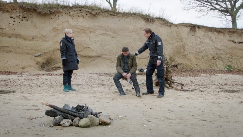 Der von der Verstorbenen getrennt in Schwerin lebende Ehemann Ingo Keller (Tom Mikulla, M.) wird zur Vernehmung nach Wismar geladen. Doch seine Ankunft verzögert sich – was ist passiert? Die Polizisten Stine (Sidsel Hindhede, l.) und Timmermann (Mathias Junge, r.) finden den völlig betrunkenen Witwer schließlich am Strand. Wo war er zur Tatzeit? Hatte er etwa Streit mit Marion Keller? – Bild: ZDF /​ Meyerbroeker